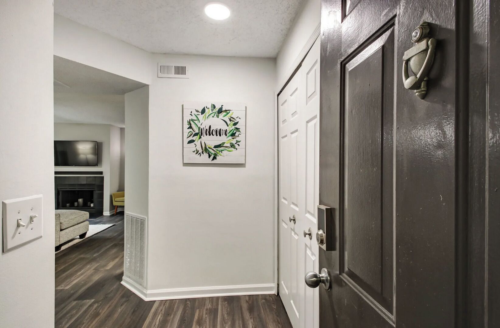 A hallway with a dark door, a welcome sign, a fireplace, and a TV in the background.