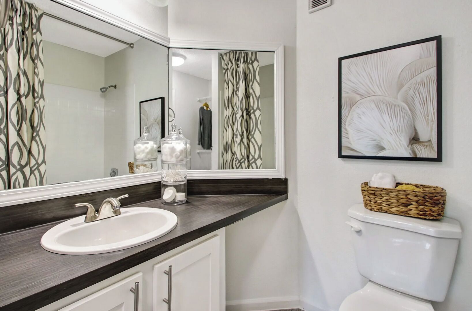 A bathroom with a white sink, dark countertop, and a large mirror. There are two glass jars on the counter, a white toilet, and a framed picture on the wall.