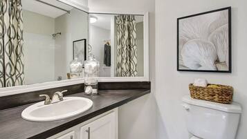 A bathroom with a white sink, dark countertop, and a large mirror. There are two glass jars on the counter, a white toilet, and a framed picture on the wall.