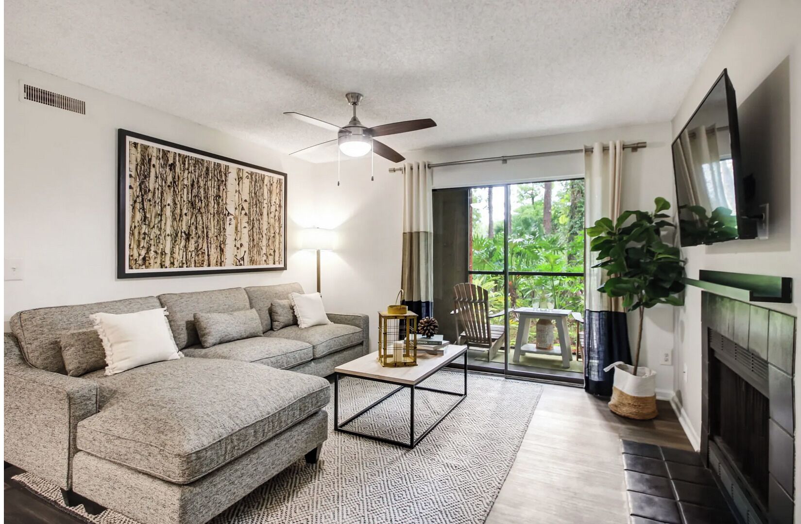 A living room with a grey sofa, a coffee table, a ceiling fan, a fireplace, and a large window with a view of greenery.