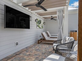 Un'area patio coperta con una TV a parete, due ventilatori a soffitto e una zona salotto con due poltrone e una panca imbottita.