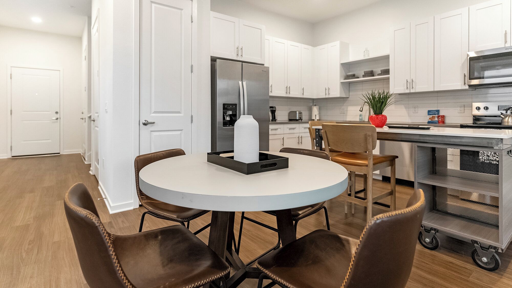 A modern kitchen with white cabinets, a stainless steel refrigerator, a microwave, and a coffee machine. There is a round table with chairs and a potted plant on the counter.