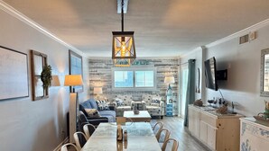 A spacious living room with a large dining table, comfortable seating, a television, and a beach-themed decor.