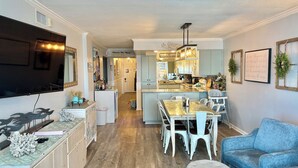 A modern kitchen with a central island, dining area with white chairs, a blue sofa, and a flat-screen TV mounted on the wall.