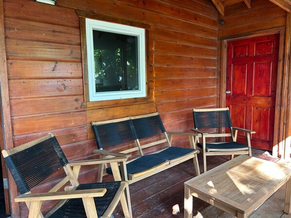 Una cabaña de madera con puerta roja, dos sillas de madera con cojines negros, una mesita y una ventana con marco blanco.