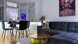 A modern living room with a dark grey sofa, a round gold coffee table, and a dining area with a white table set for two. - Stylish Apartment in Buckhurst Hill (London)