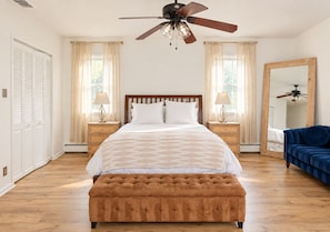 A bedroom with a bed, a ceiling fan, a large mirror, a blue sofa, and a wooden floor. - Heated Pool, Gym & Game Room, 5 Min to Beach (Hampton Bays)