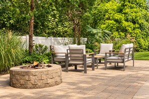 An outdoor patio with a stone fire pit, two grey lounge chairs, and a wooden table. - Heated Pool, Gym & Game Room, 5 Min to Beach (Hampton Bays)
