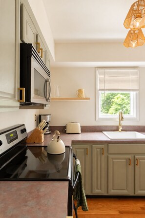 A modern kitchen with a stove, microwave, and toaster. There is a window with blinds and a hanging light fixture. - Heated Pool, Gym & Game Room, 5 Min to Beach (Hampton Bays)