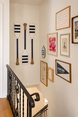 A staircase with a black railing and a wooden handrail, leading to a landing with wall-mounted artwork including a framed picture of a lighthouse and a sign that reads 'HAMPTONS surf house'. - Heated Pool, Gym & Game Room, 5 Min to Beach (Hampton Bays)