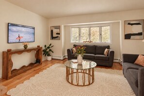 A living room with a wooden console table, a glass coffee table, a grey sofa, and a large window with a view of trees.