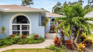 A house with a light blue exterior, large arched windows, and a well-maintained garden with various plants and a palm tree. - Heated Pool & Hot Tub | 4BR Sarasota Home – Pets OK (Sarasota)
