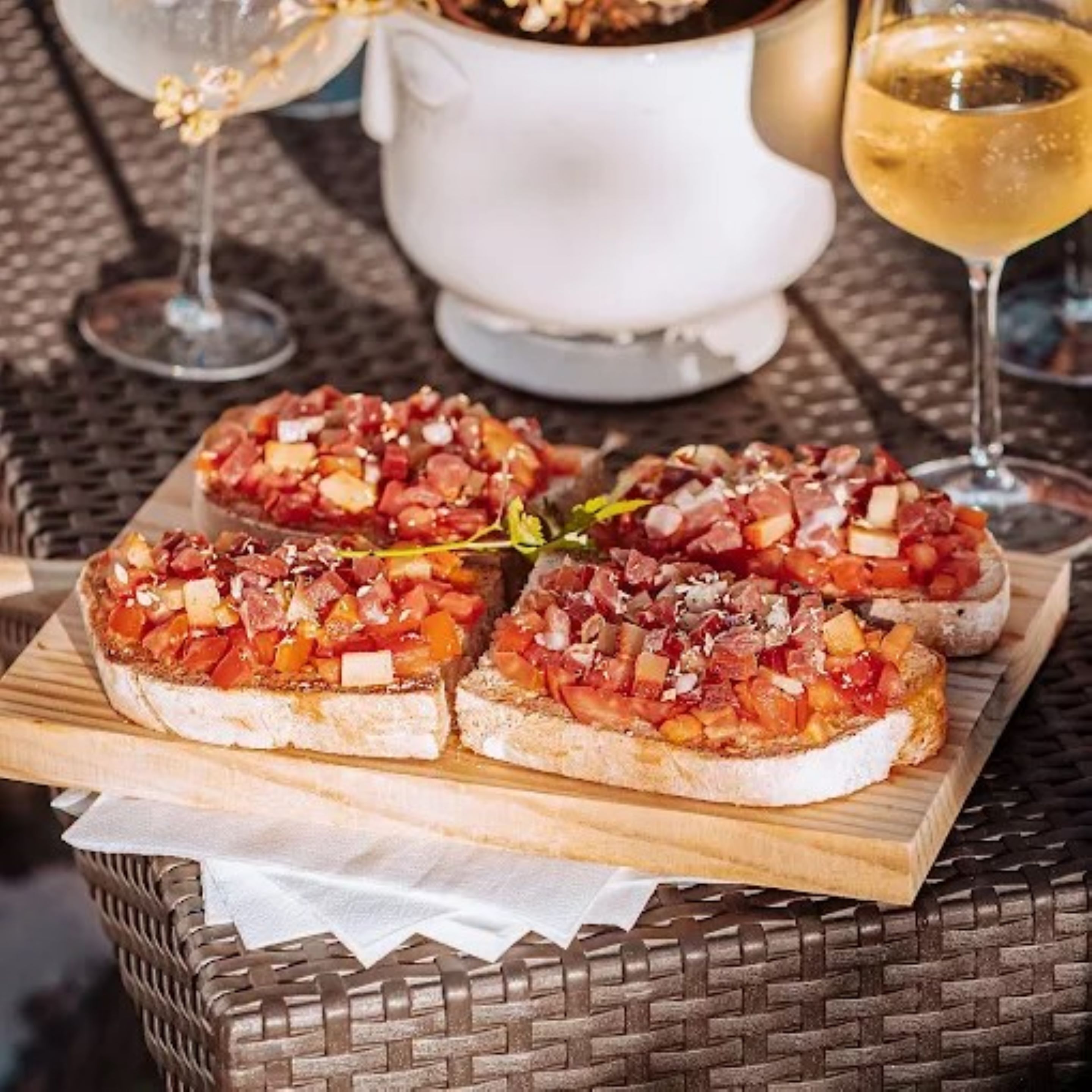 Four pieces of bruschetta on a wooden board, a white pitcher, and a glass of white wine on a wicker table.