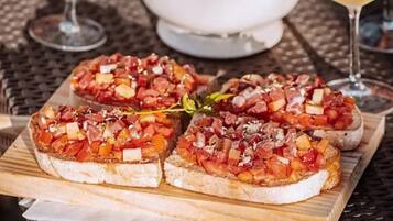 Four pieces of bruschetta on a wooden board, a white pitcher, and a glass of white wine on a wicker table.