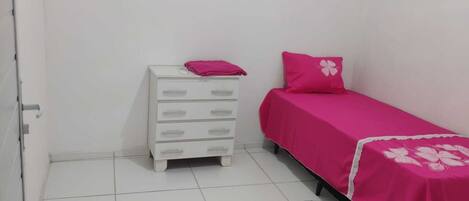 A small, clean room with a white dresser, a single bed with a pink bedspread, and a white door.