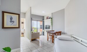 Una habitación moderna y bien iluminada con cama, una mesa de comedor con sillas, una pequeña cocina y un ventanal que ofrece vistas al exterior.