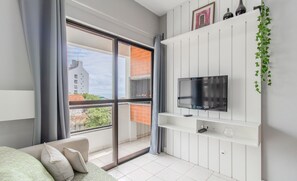 Una sala de estar moderna con un amplio ventanal corredera que da a un balcón, un televisor de pantalla plana montado en una pared blanca y un pequeño estante con objetos decorativos.