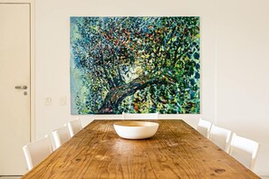 Un comedor con una mesa de madera y sillas blancas, un cuadro abstracto grande en la pared, y una puerta con manija.