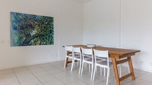 A dining area with a wooden table and white chairs, a painting on the wall, and a tiled floor.