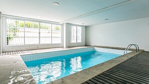 A swimming pool with tiled walls and a wooden deck, large windows with white frames, and a ceiling with recessed lighting.