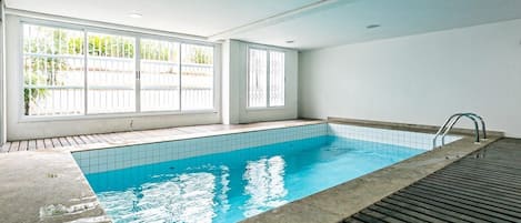 Una piscina con paredes de azulejos y un deck de madera, ventanas amplias con marcos blancos e iluminación empotrada en el techo.