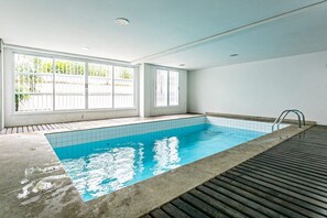 Una piscina con paredes de azulejos y un muelle de madera, ventanas amplias con marcos blancos e iluminación empotrada en el techo.