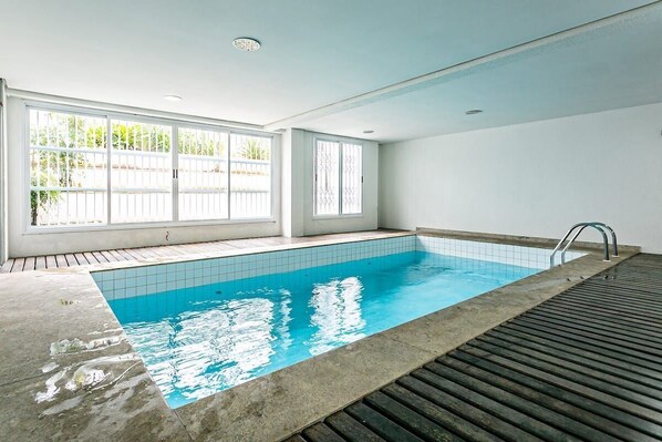 Una piscina con paredes de azulejos y un deck de madera, ventanas amplias con marcos blancos e iluminación empotrada en el techo.