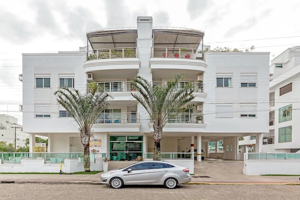 Un edificio de varios pisos con balcones, un coche estacionado al frente y palmeras.