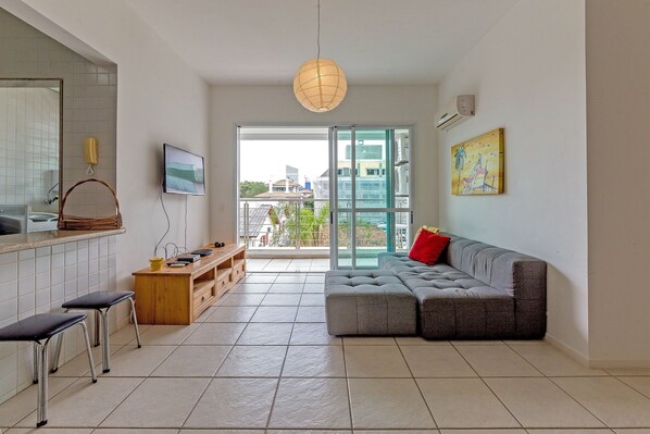 A modern living room with a grey sectional sofa, a wooden TV stand, and a large window offering a view of buildings and greenery.