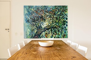 Un comedor con una mesa de madera y sillas blancas, un cuadro abstracto grande en la pared, y una puerta con manija.