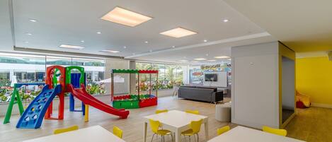 A modern indoor play area with slides and climbing structures, surrounded by yellow chairs and tables.