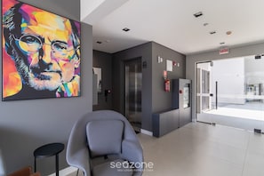 Un moderno vestíbulo de hotel con un cuadro abstracto de gran tamaño, una recepción, un ascensor y una puerta de cristal que da al exterior.