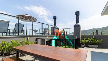 A rooftop terrace with a wooden table, chairs, a children's play area, and a view of the surrounding landscape.