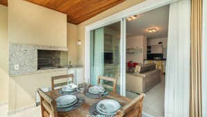 A dining area with a wooden table set for four, a built-in oven, and a sliding glass door leading to a living area. - Beachfront Apartment w/ View Beach Santinho Asr204 (Florianópolis)