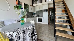 A modern kitchen with white cabinets and a black countertop, a dining area with a patterned tablecloth, and a wooden staircase. - Le Mezmarine - Front de Mer (Bray-Dunes)