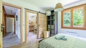 A bedroom with a bed, a wooden wardrobe, a bathroom with a sink and mirror, and a view of greenery through a window.
