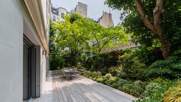 Une terrasse en bois avec une table et des chaises, entourée d’une végétation luxuriante et d’un arbre.
