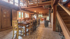 A spacious dining area with wooden tables and chairs, a high ceiling with exposed beams, and a view of the kitchen in the background.