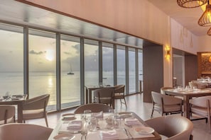 A dining area with tables set for two, large windows offering a view of the sea and boats, and a modern interior design. - Royalton Vessence Barbados Adult-Oriented, an Autograph Collection All-Inclusive Resort – Now Open (Holetown)