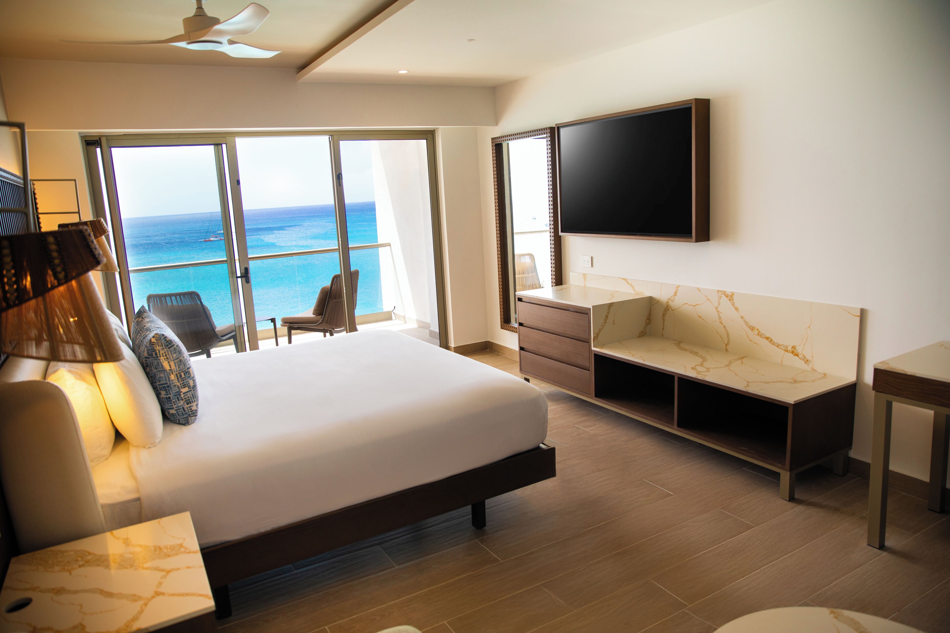 A modern hotel room with a large bed, a flat-screen TV, a desk, and a view of the ocean.