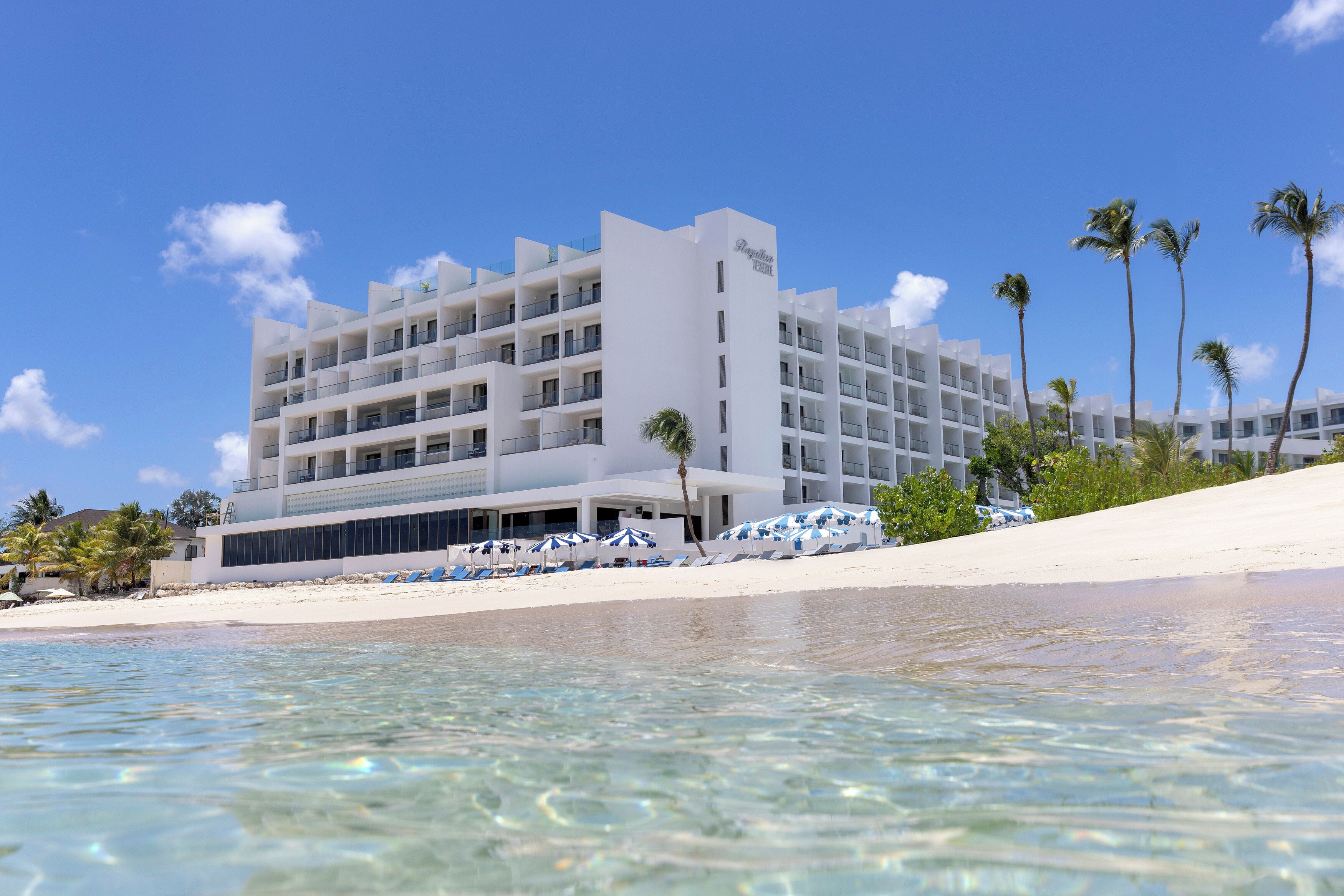 Un hotel frente a la playa con varios balcones, una playa de arena y palmeras.
