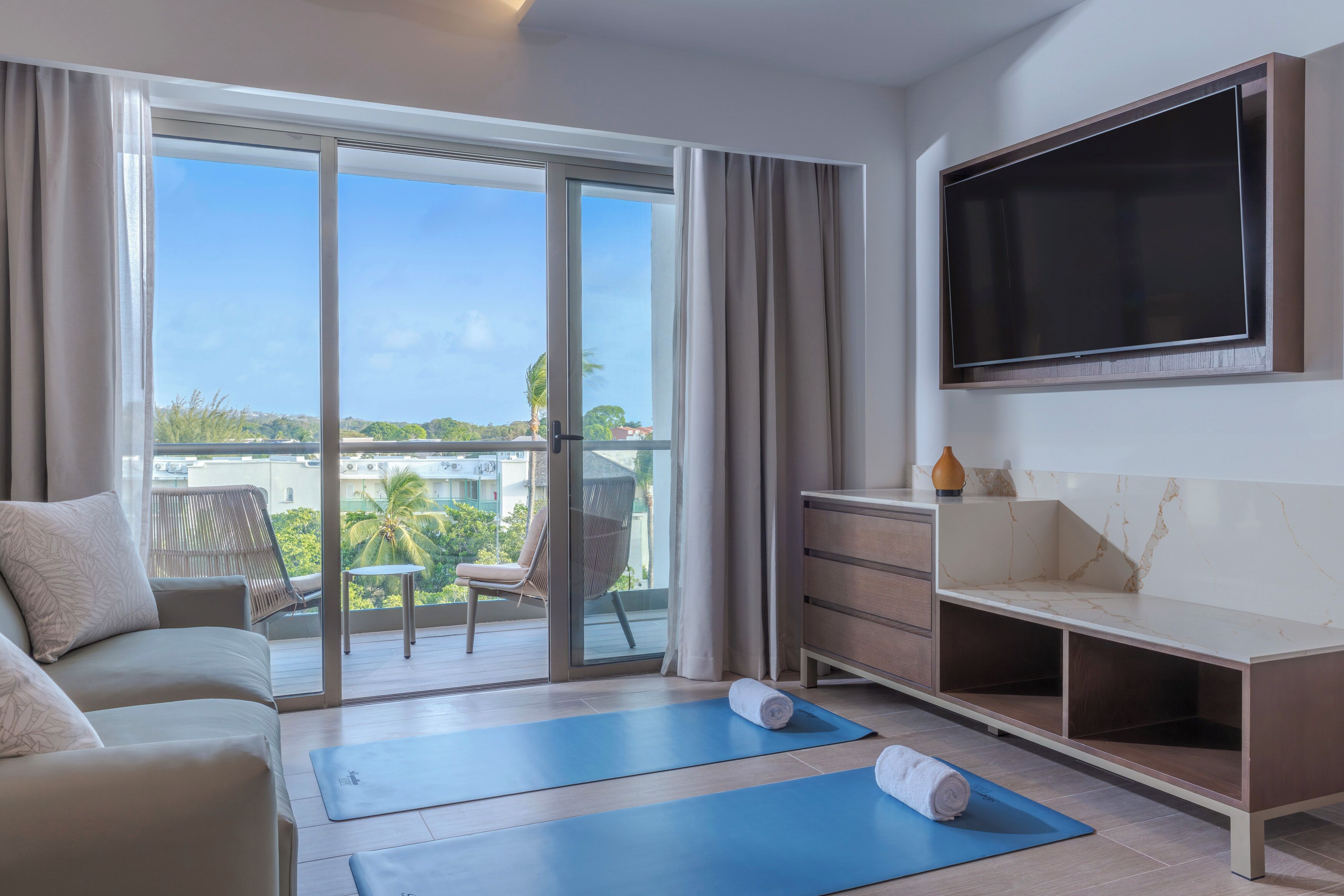 A hotel room with a sofa, a TV mounted on the wall, a balcony with a view, and exercise mats on the floor.