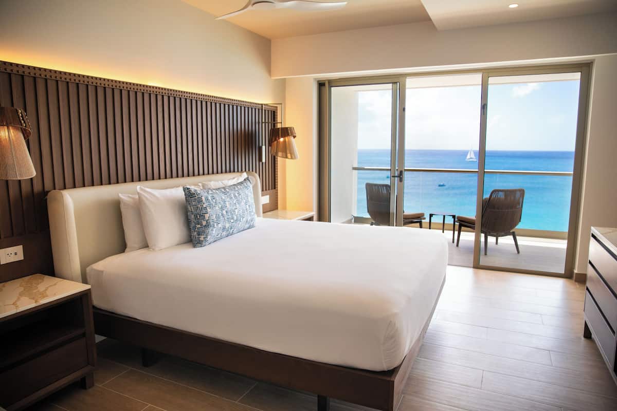 A hotel room with a large bed, a nightstand, a lamp, a sliding glass door to a balcony with a view of the sea.