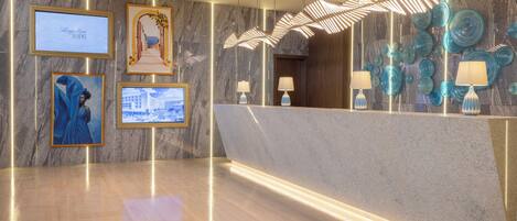 A modern hotel lobby with marble walls, framed artwork, and a reception desk with decorative lamps.