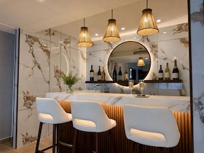 Een moderne bar met marmeren wanden, drie witte barkrukken, een ronde spiegel en hangende hanglampen.