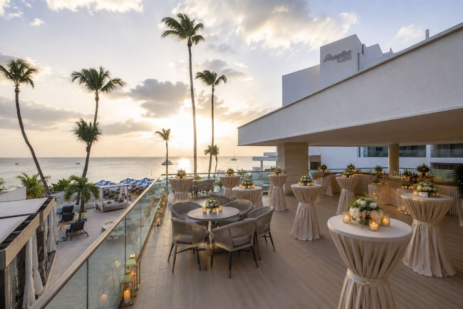 A rooftop terrace with tables and chairs, palm trees, and a view of the ocean.