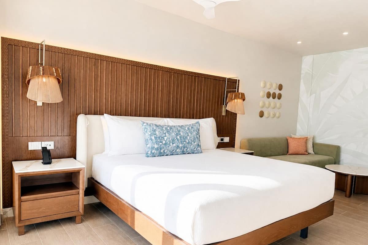 A modern hotel room with a wooden headboard, a large bed with white bedding, a bedside table with a lamp, a green sofa, and a small round table.