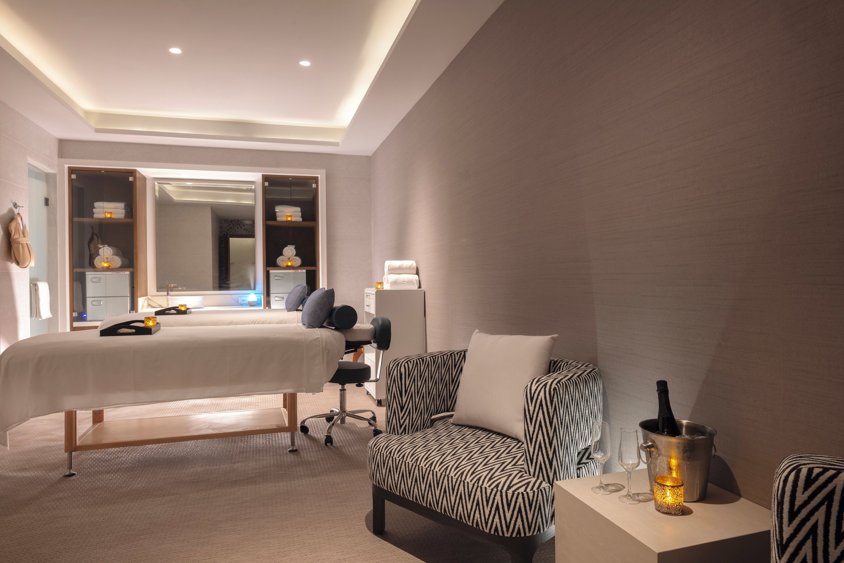 A hotel room with a bed, a desk, a chair, a bedside table with a bottle, and a nightstand.
