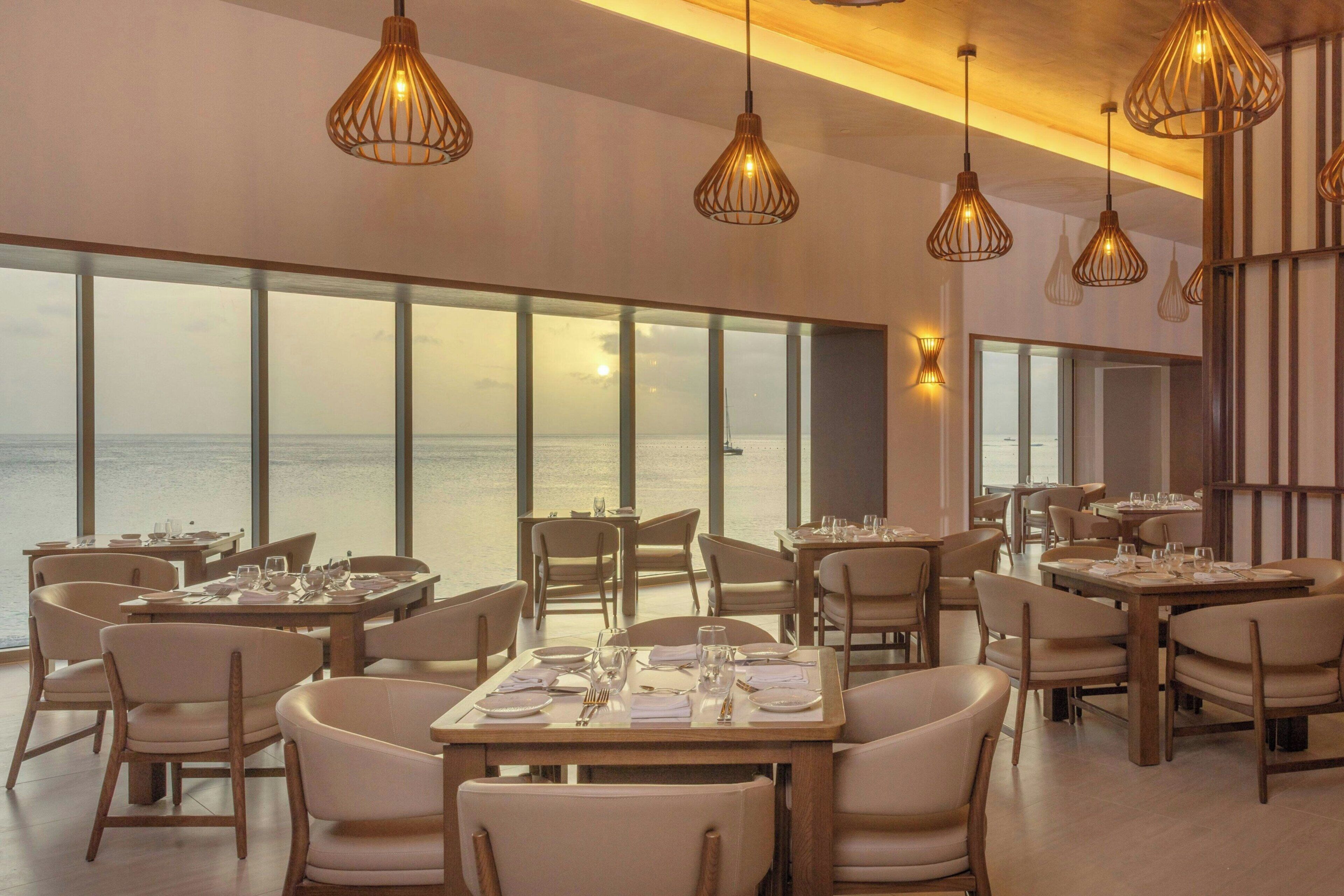 Un restaurante con mesas dispuestas para comer, amplias ventanas con vistas al mar y lámparas colgantes.