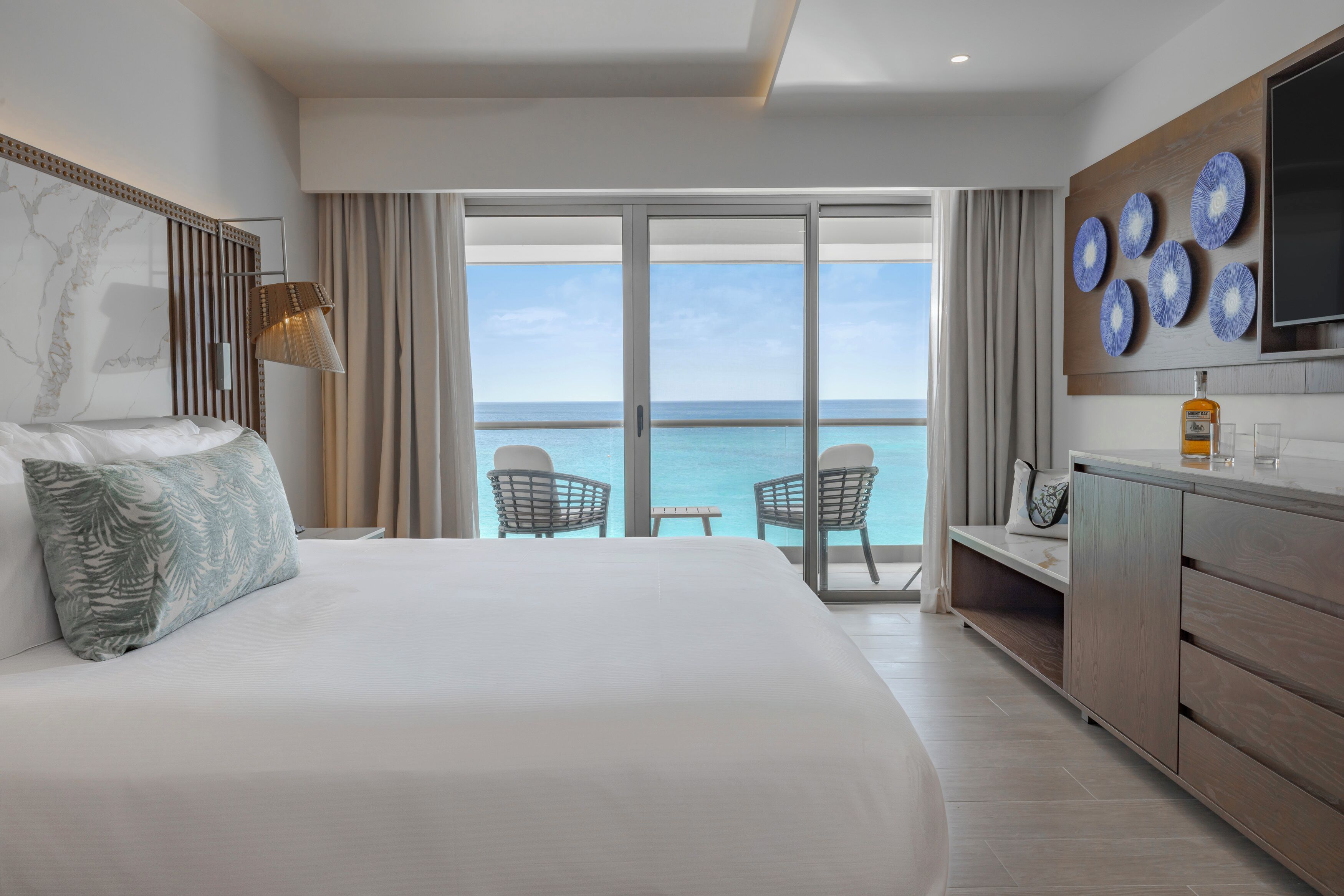 Habitación de hotel con cama, televisor, minibar, balcón con vista al mar y zona de comedor con sillas.
